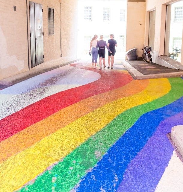 Kolme henkilöä kävelee porttiholvissa. Maahan on maalattu sateenkaarimaalaus.